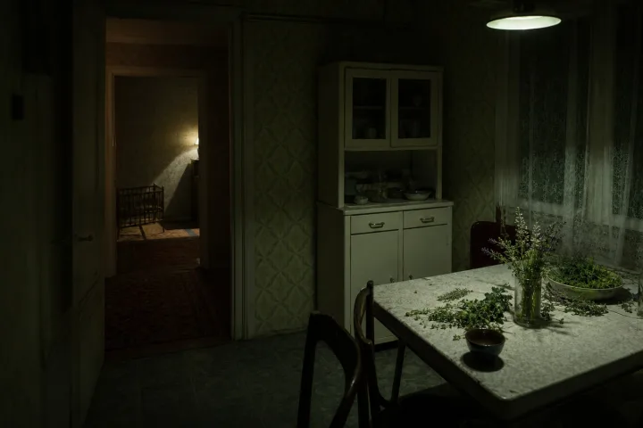 Cucina notturna anni Sessanta con erbe sparse sul tavolo e una culla in ombra nella stanza vicina
